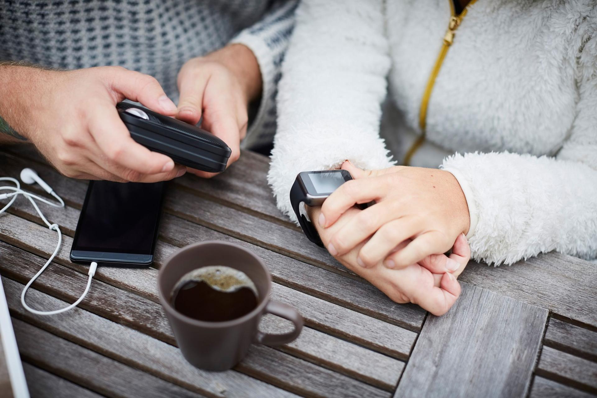 En kvinna med en smart klocka och en man med en mobiltelefon sitter vid ett trädgårsbord. En kaffekopp står på bordet.