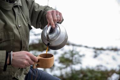 En person i en grön jacka står utomhus på vintern och häller upp kaffe i ett träkåsa ur en kaffepanna.